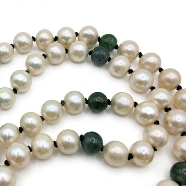 A close-up of a beaded necklace featuring round off-white freshwater pearls separated by small black beads, accented with a few dark green faceted beads, set against a plain white background.