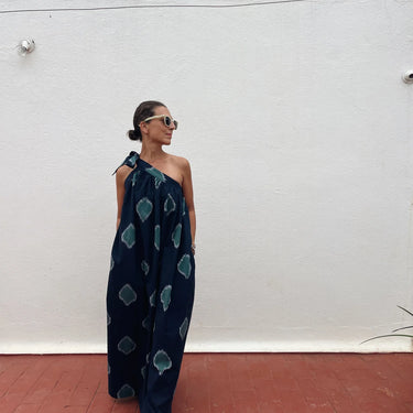 A woman wearing a navy blue one-shoulder jumpsuit with a teal ikat pattern, standing against a white wall on a red brick floor.