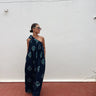 A woman wearing a navy blue one-shoulder jumpsuit with a teal ikat pattern, standing against a white wall on a red brick floor.